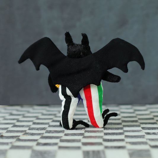 Hand-sewn black bat plush with wide fabric wings and multicolored striped pants, shown from behind on a checkered surface with a dark background