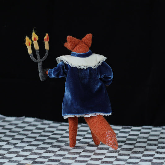Back view of a handmade fox doll wearing a blue velvet dress with a white lace collar, holding a three-flame felt candelabra while standing on a black-and-white checkered floor against a dark background