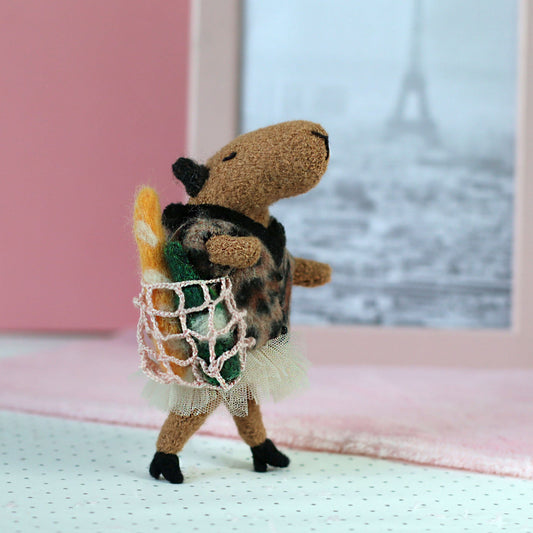 Handmade miniature capybara doll standing with a tiny mesh shopping bag holding a felt baguette and bottle, wearing a leopard-print sweater and tulle skirt, with a pink backdrop and Eiffel Tower photo in the background
