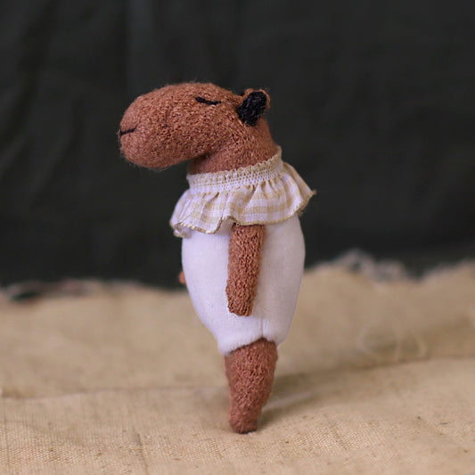 Side view of a handmade miniature capybara doll in a white romper with a ruffled checked collar, standing on beige fabric with a peaceful closed-eye expression