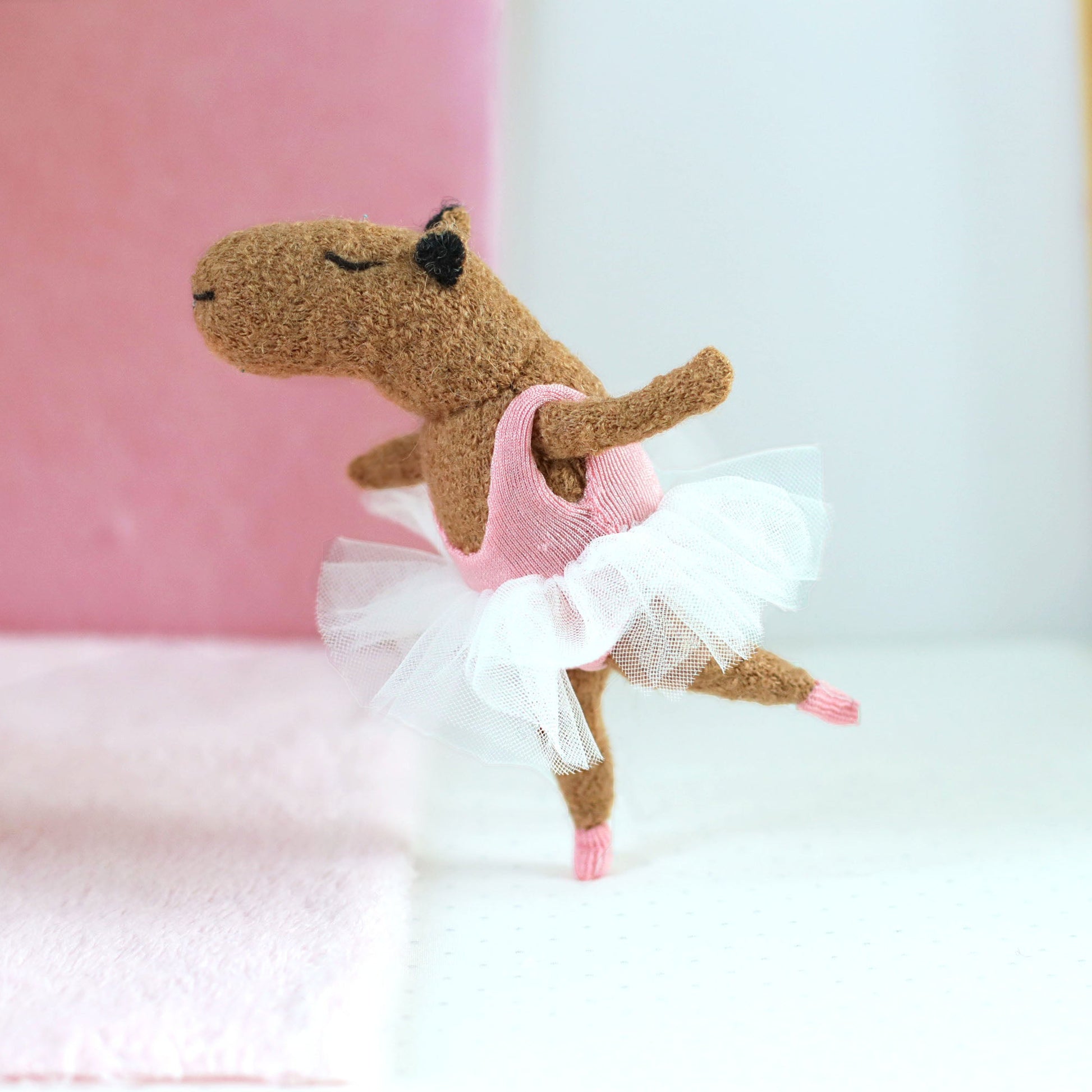 Handmade capybara ballerina doll posed in an arabesque, wearing a pink leotard and white tulle tutu, standing on one leg in a soft pink and white background