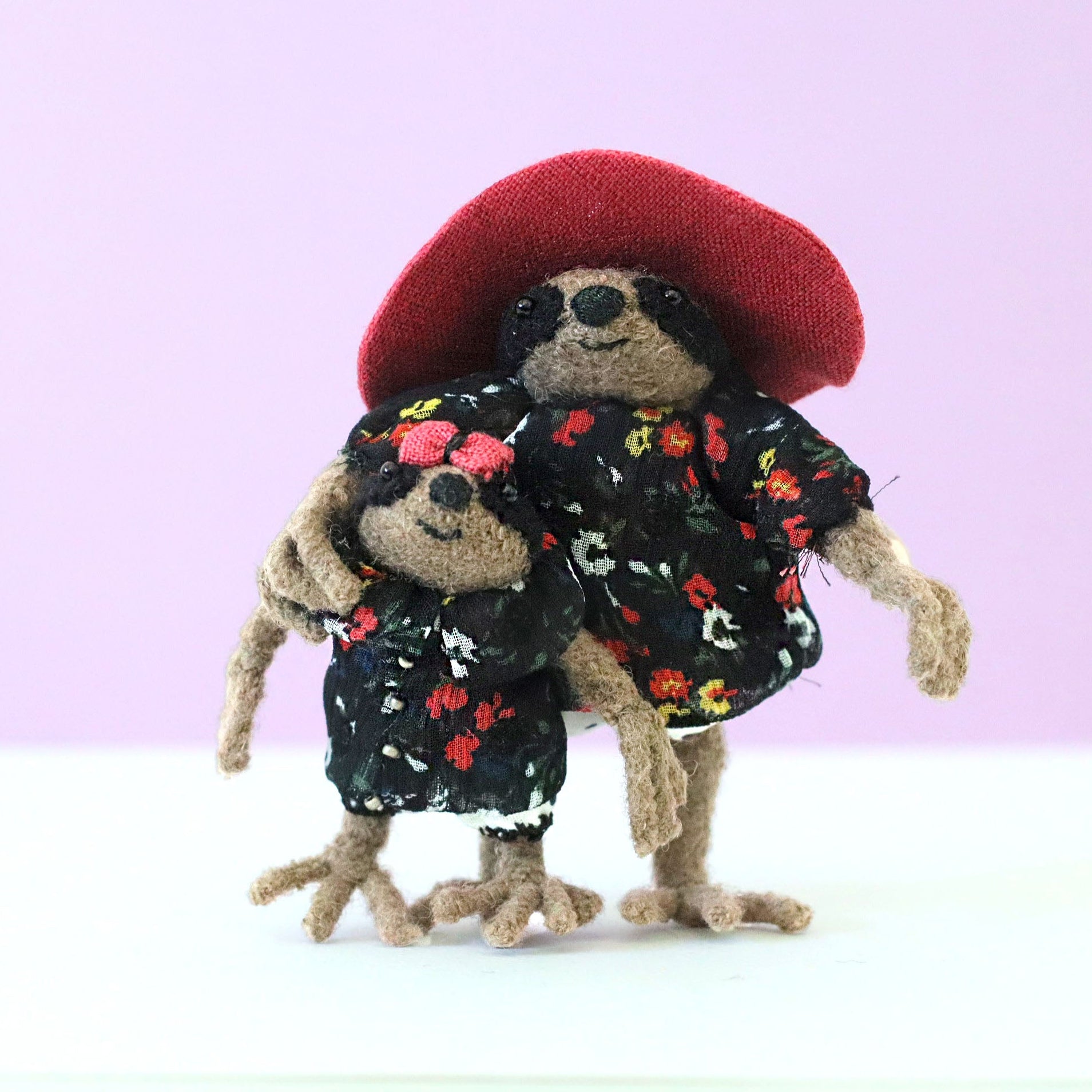 Mama and baby sloth plush dolls hugging in matching floral dresses, with the mother in a wide red hat, against a soft pink background
