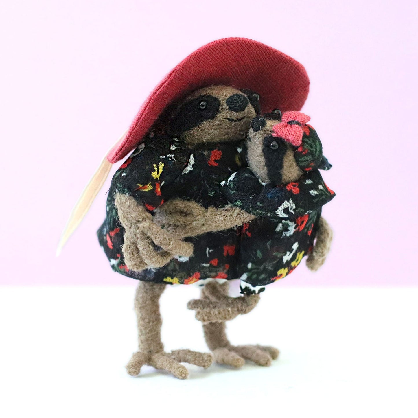 Handmade mama sloth doll in a pink sun hat holding her baby, both dressed in matching black floral dresses, posed against a soft pastel background