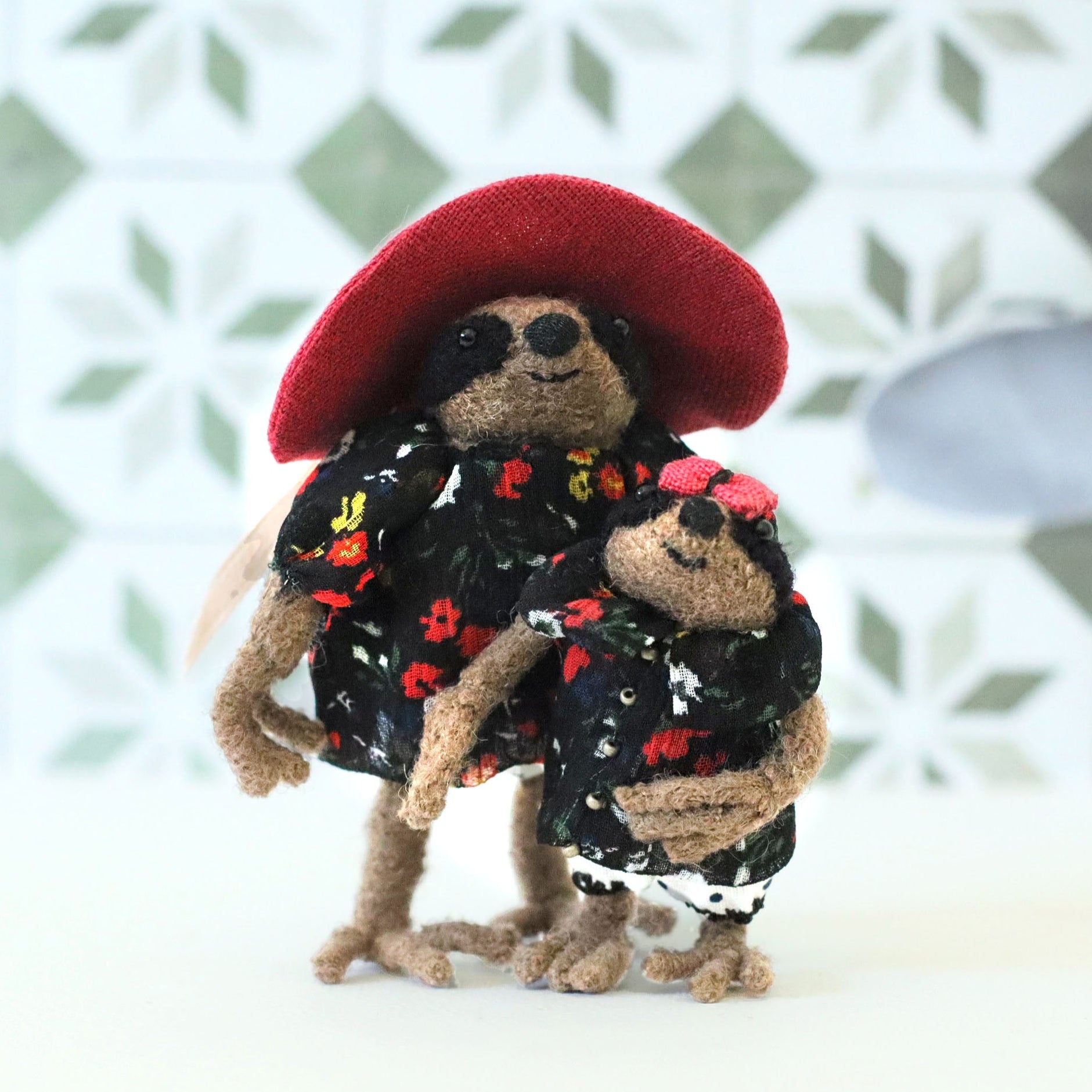 Handcrafted mama sloth doll wearing a red sun hat standing beside her baby sloth, both dressed in matching black floral outfits, posed against a patterned tile background