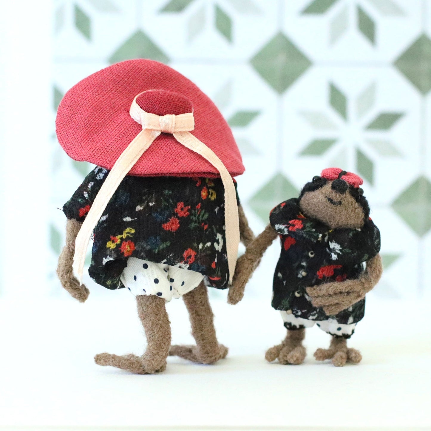 Back view of a handmade mama sloth doll in a floral dress and large pink sun hat, holding hands with a baby sloth doll in a matching floral dress and tiny bow