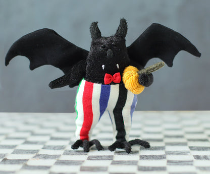 Hand-sewn black bat plush with fabric wings, a red bow tie, colorful striped pants, and a tiny orange pumpkin in its hand, standing on a checkered surface