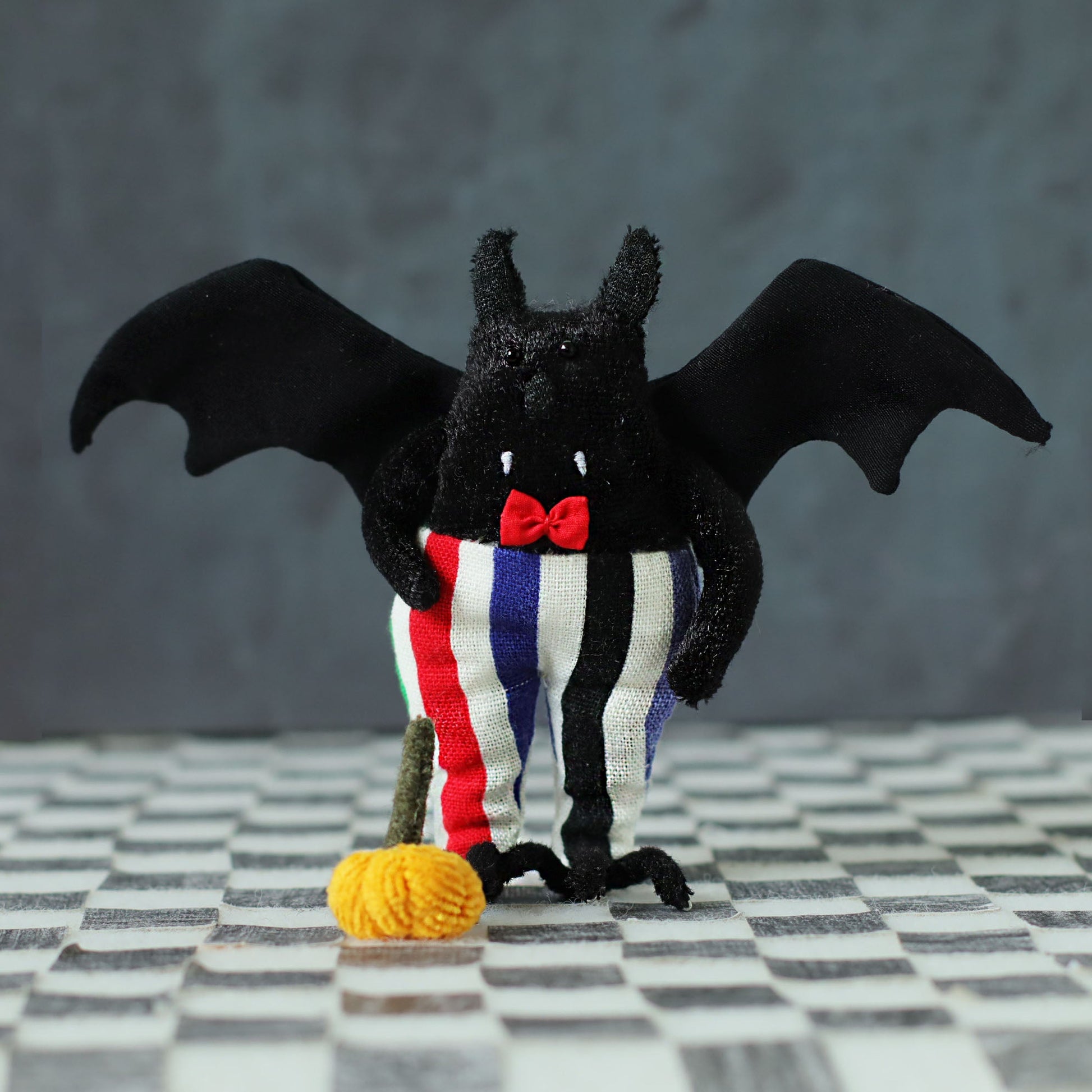 Hand-sewn black bat plush with fabric wings, a red bow tie, and colorful striped pants, standing on a checkered surface with a tiny orange pumpkin in front of it