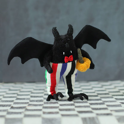 Soft handmade bat plush with black wings, a red bow tie, and colorful striped trousers, holding a tiny orange pumpkin while standing on a checkered surface