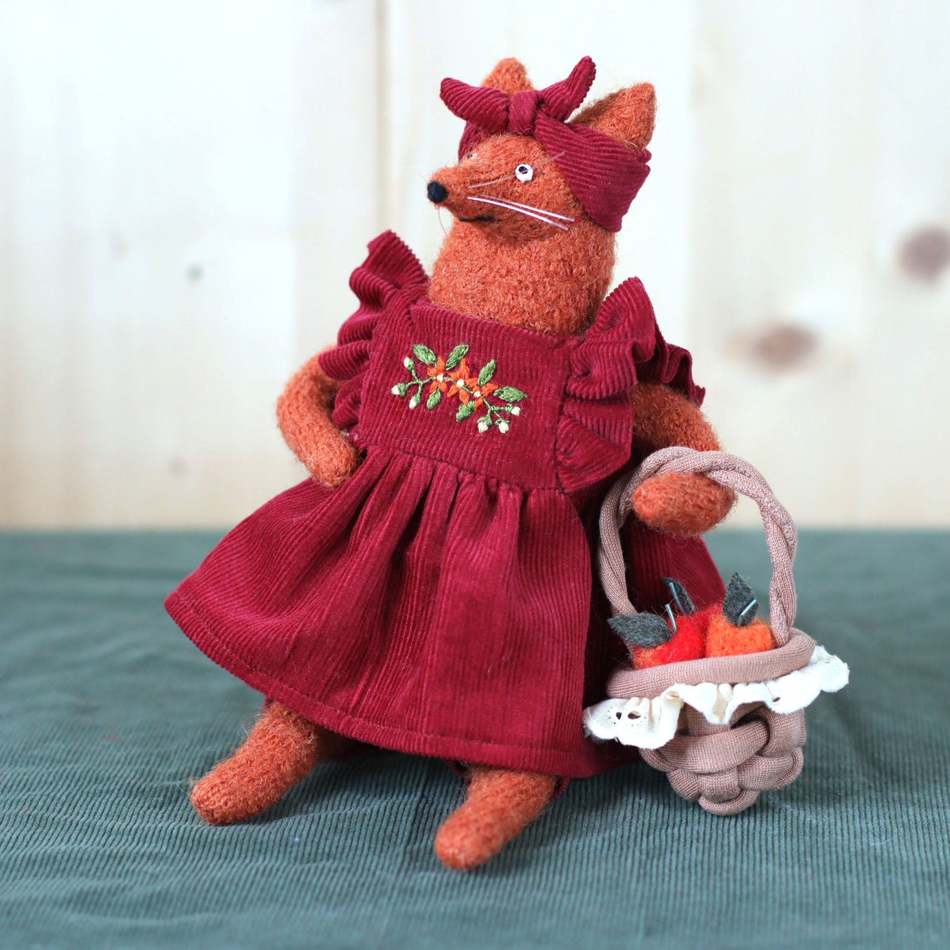 Handmade fox doll wearing a red embroidered dress and matching headband, sitting and holding a small basket with felt apples