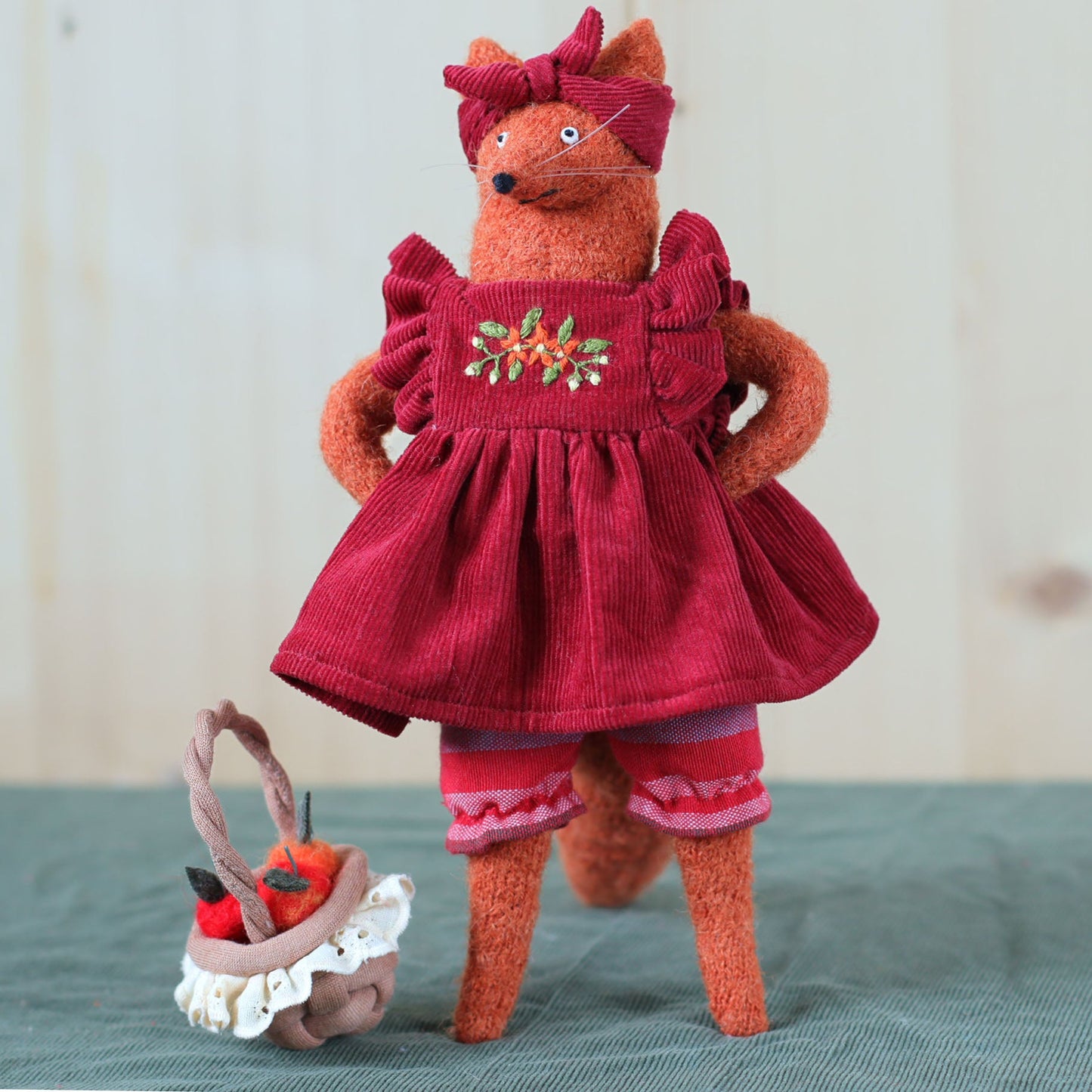 Handmade fox doll in a red embroidered dress, standing beside a small basket filled with felted apples