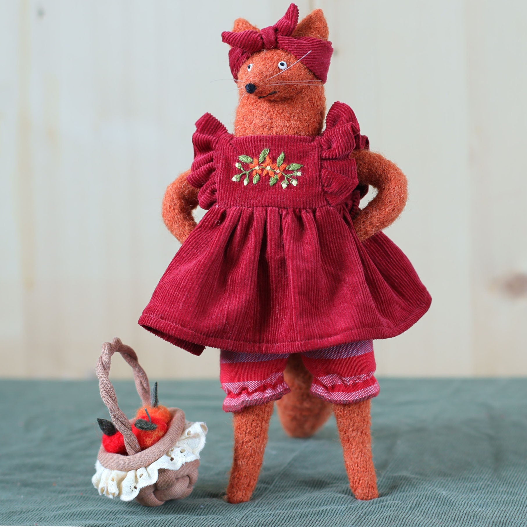 Handmade fox doll standing upright in a red embroidered dress and matching headband, with a small basket of felt apples placed beside her