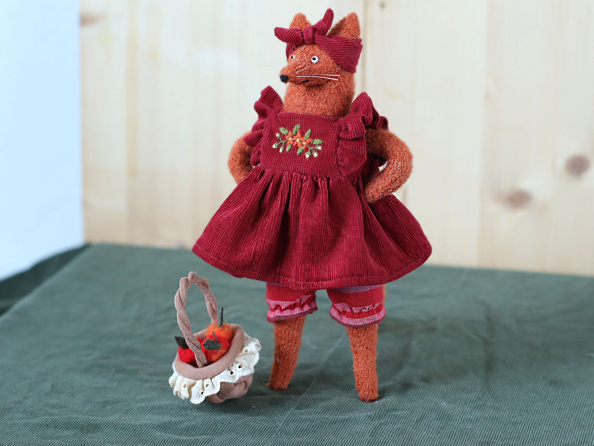 Handmade fox doll standing on a green surface, dressed in a red embroidered dress with a matching headband, next to a small basket filled with felt apples