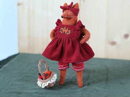 Handmade fox doll standing on a green surface, dressed in a red embroidered dress with a matching headband, next to a small basket filled with felt apples