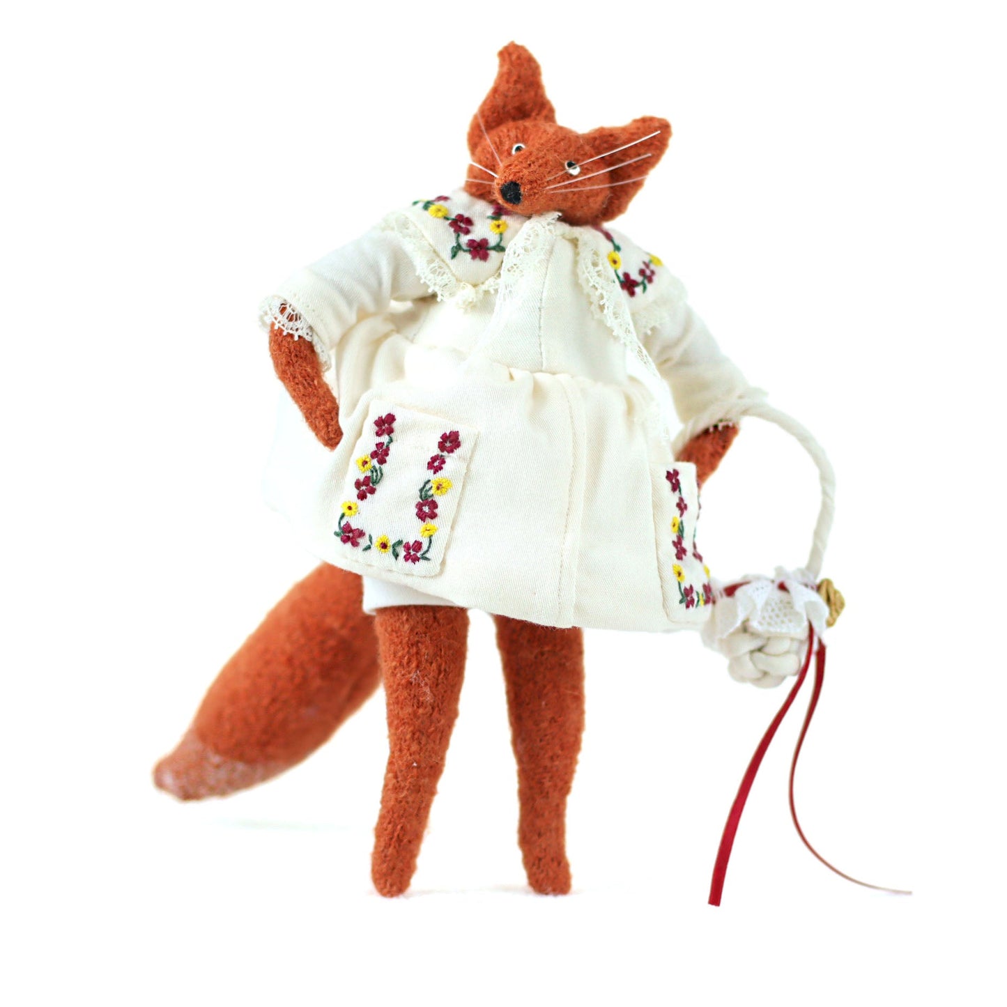 Handmade fox doll wearing an embroidered white dress, standing upright and holding a small woven bag with a red ribbon.
