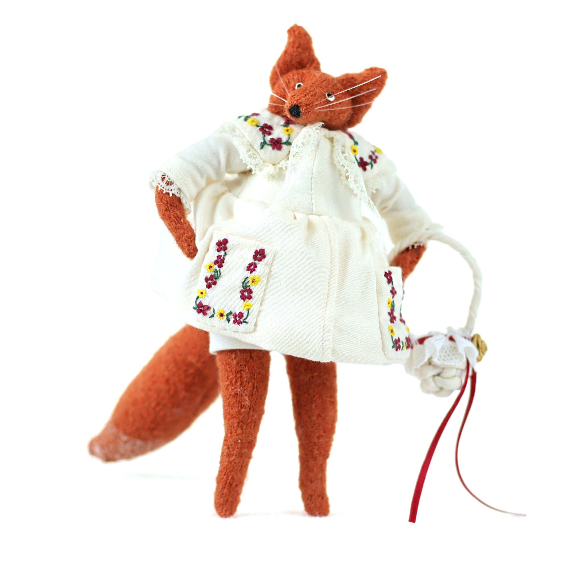 Handmade fox doll wearing an embroidered white dress, standing upright and holding a small woven bag with a red ribbon.

