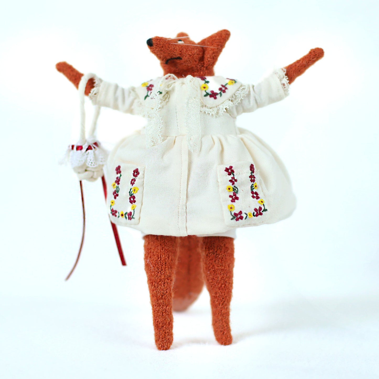 Handmade fox doll wearing an embroidered white dress with colorful floral details, standing upright with arms raised and holding a small white bag with red ribbons
