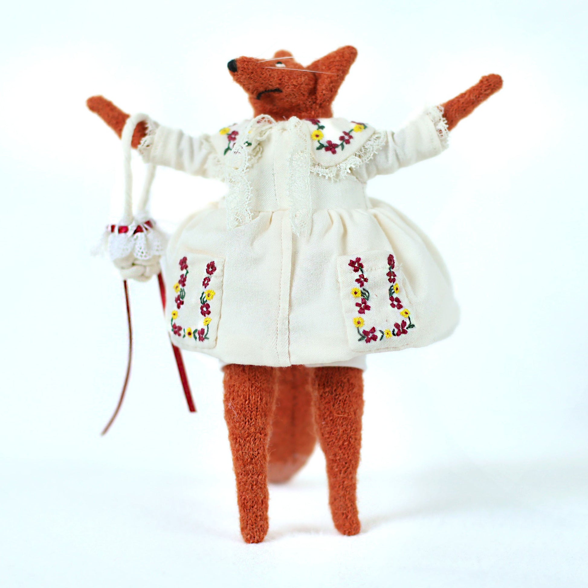 Handmade fox doll wearing an embroidered white dress with colorful floral details, standing upright with arms raised and holding a small white bag with red ribbons