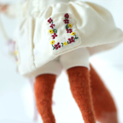 Close-up of a handmade fox doll wearing a cream dress with detailed floral embroidery on the pocket, showing the lower part of the outfit and the doll’s orange legs.