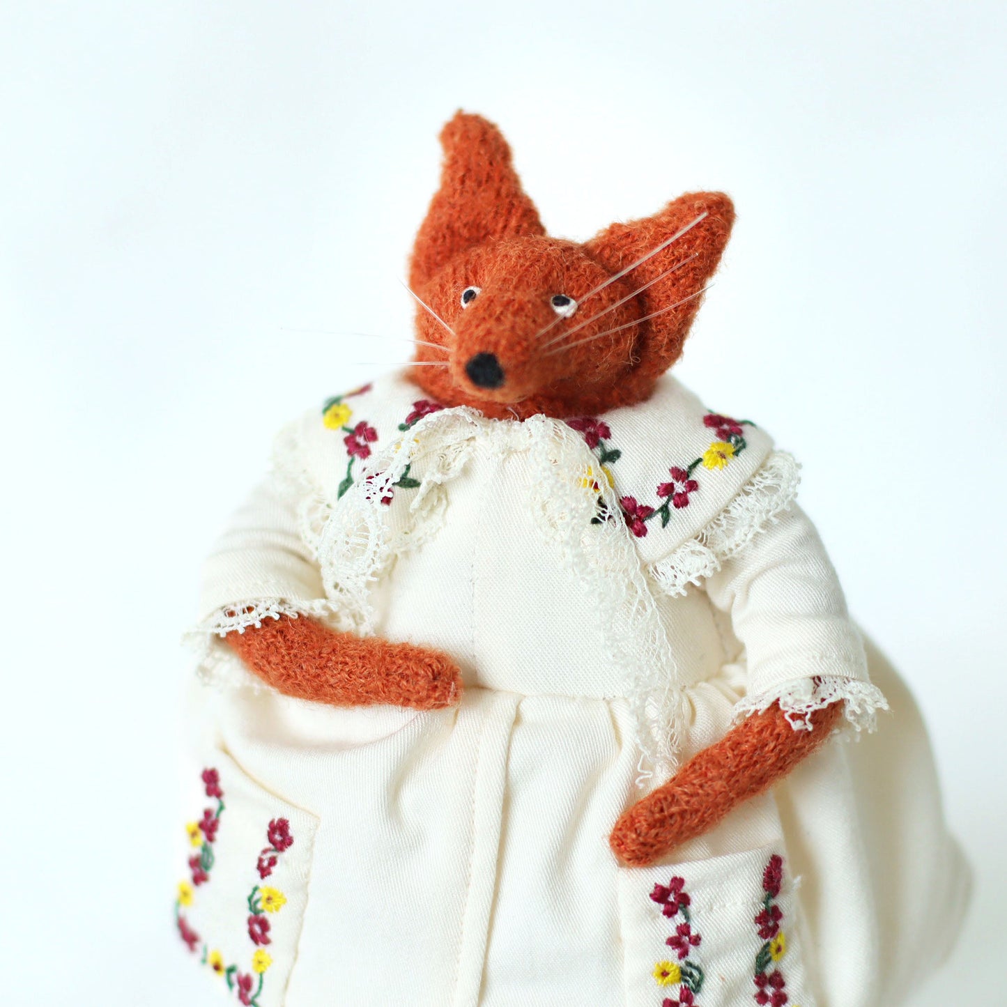 Handmade fox doll wearing a cream dress with detailed floral embroidery on the collar and pockets, shown in a close-up pose with one hand in the pocket