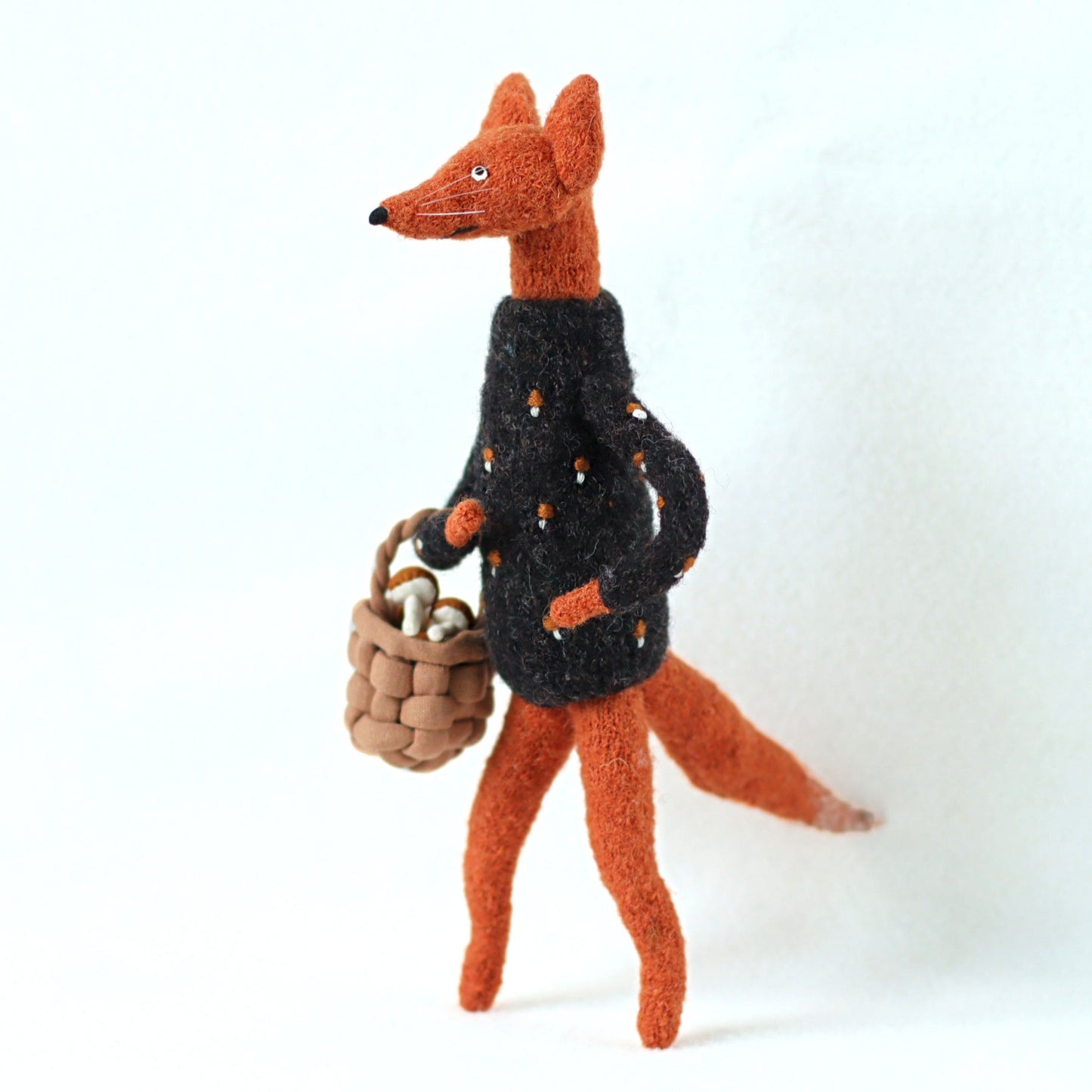 Small handmade fox doll made of wool-blend fabric, dressed in a dark embroidered sweater and carrying a tiny woven basket