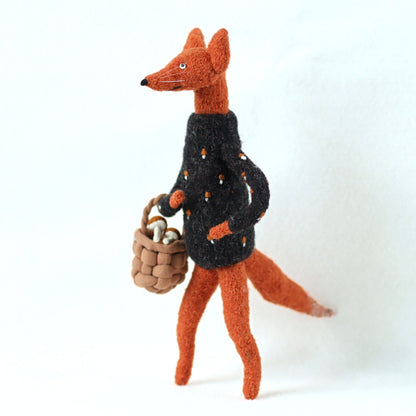 Small handmade fox doll made of wool-blend fabric, dressed in a dark embroidered sweater and carrying a tiny woven basket