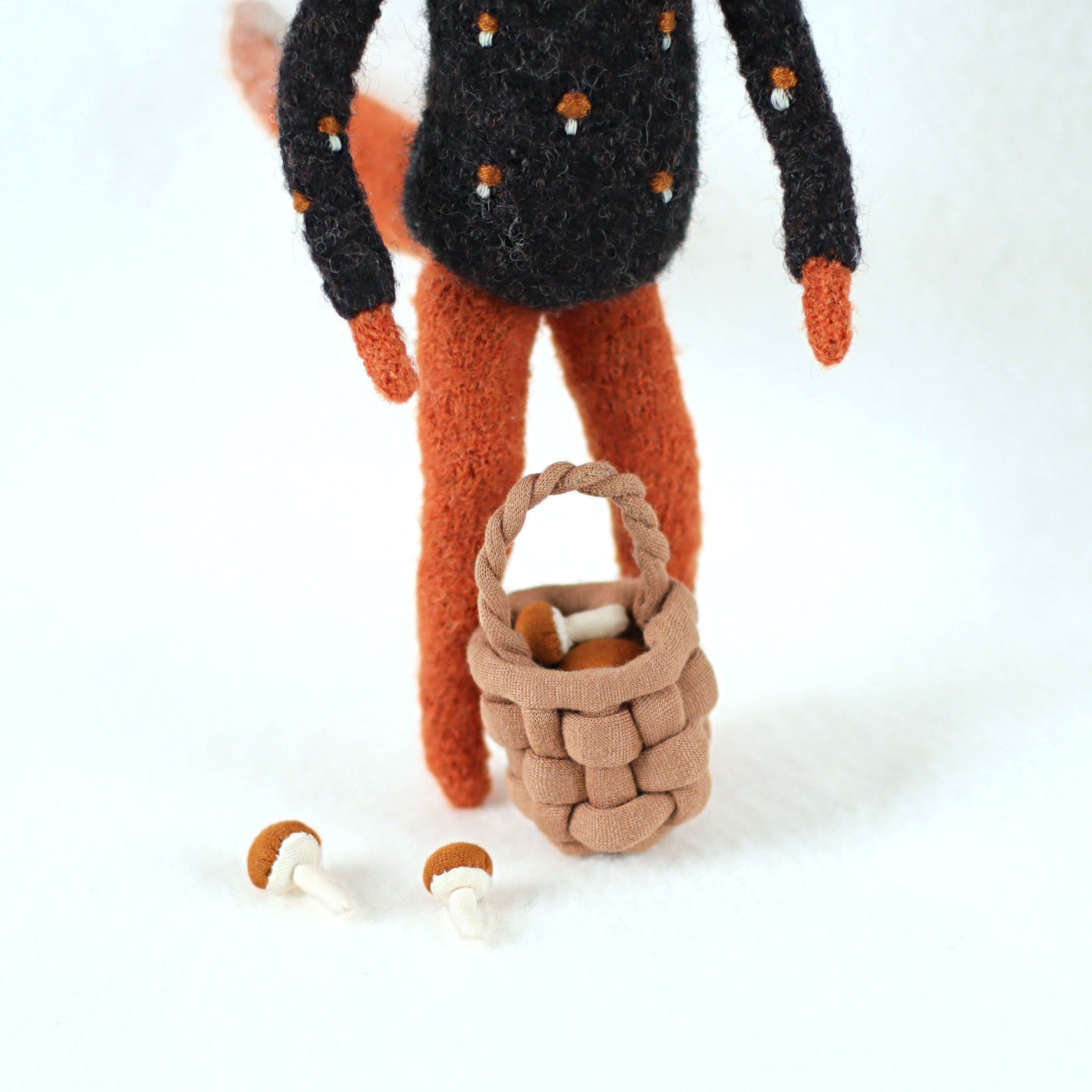 Close-up of a handmade fox figurine's legs with a small woven basket and two tiny mushrooms on the ground
