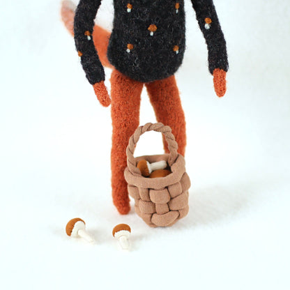 Close-up of a handmade fox figurine's legs with a small woven basket and two tiny mushrooms on the ground