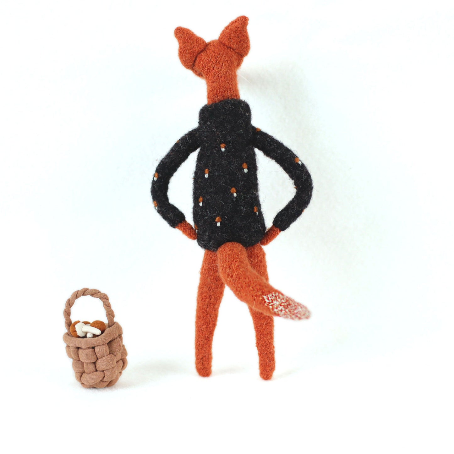 Handmade fox figurine seen from behind, wearing a dark embroidered sweater, standing beside a small woven basket filled with tiny mushrooms
