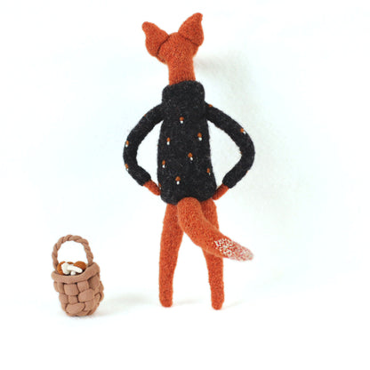 Handmade fox figurine seen from behind, wearing a dark embroidered sweater, standing beside a small woven basket filled with tiny mushrooms