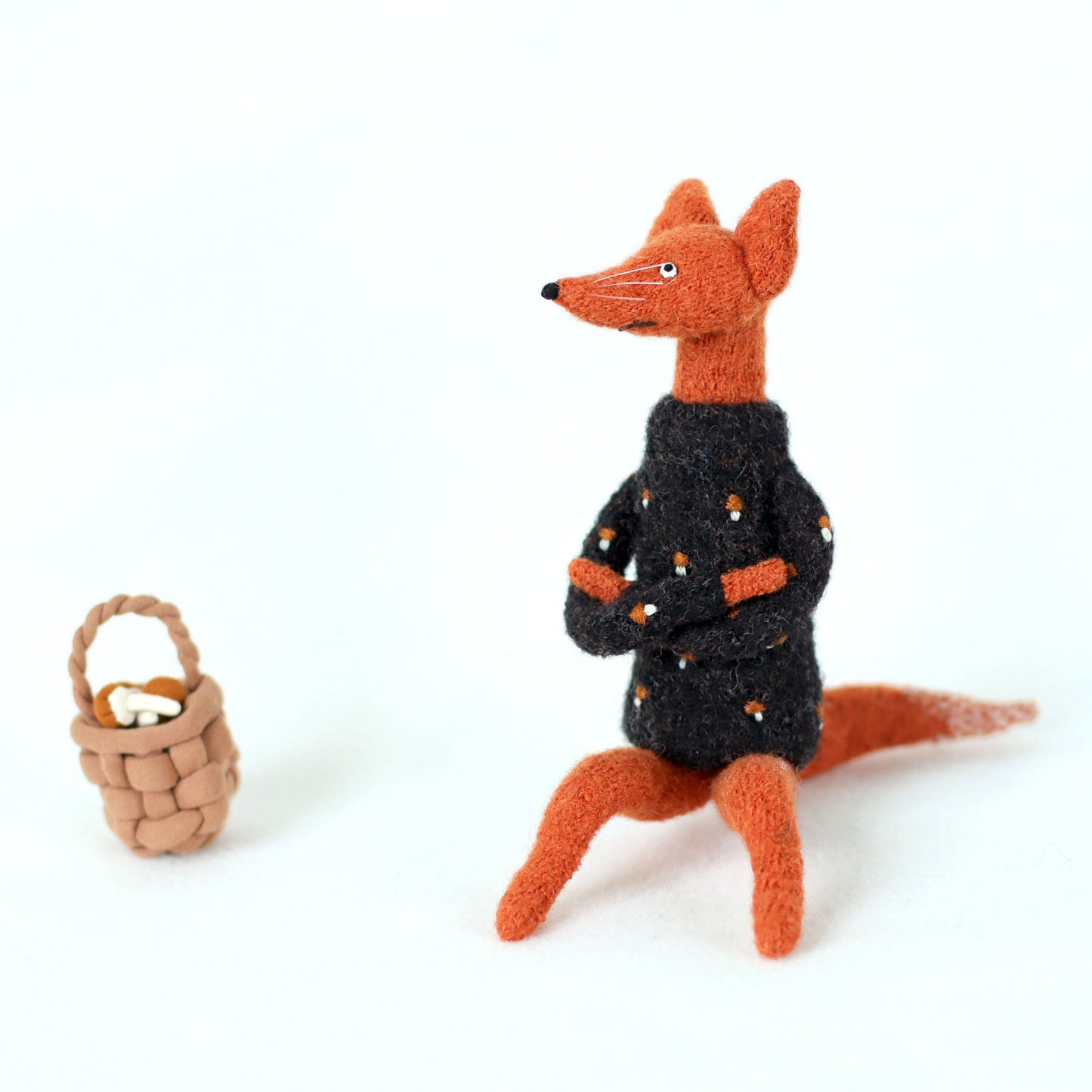 Handcrafted orange fox figurine in a black embroidered sweater, sitting with arms crossed next to a small woven basket filled with tiny mushrooms