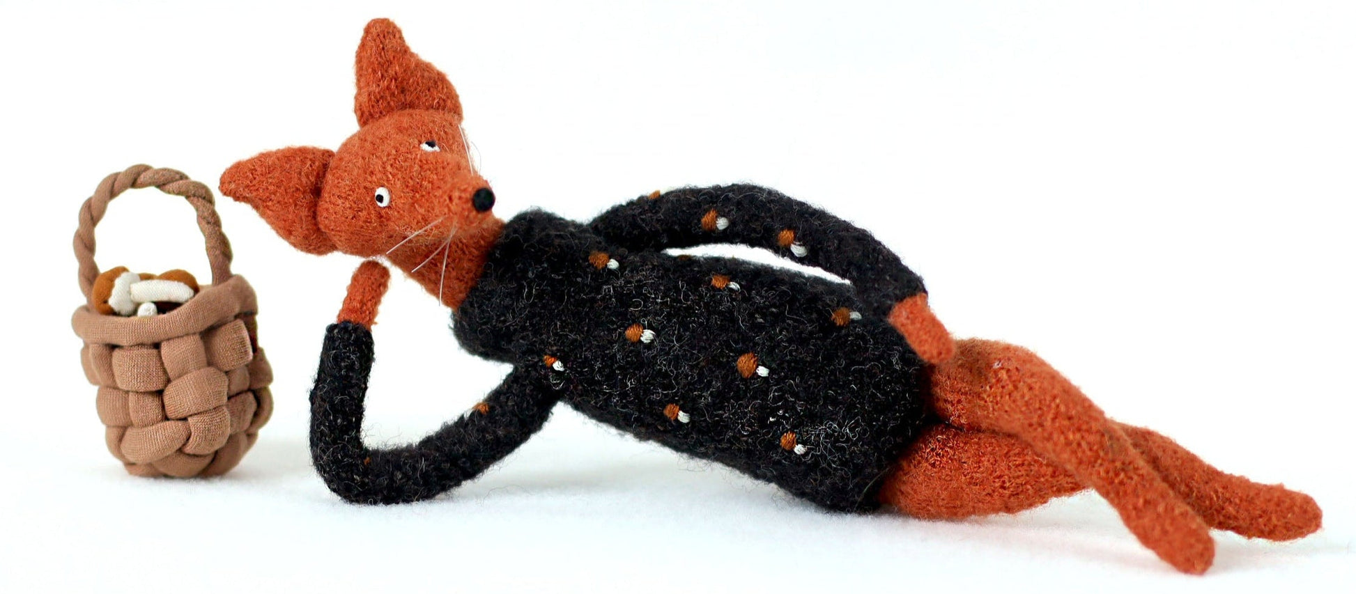 Handcrafted orange fox figurine in a black embroidered sweater lying on its side next to a tiny woven mushroom basket