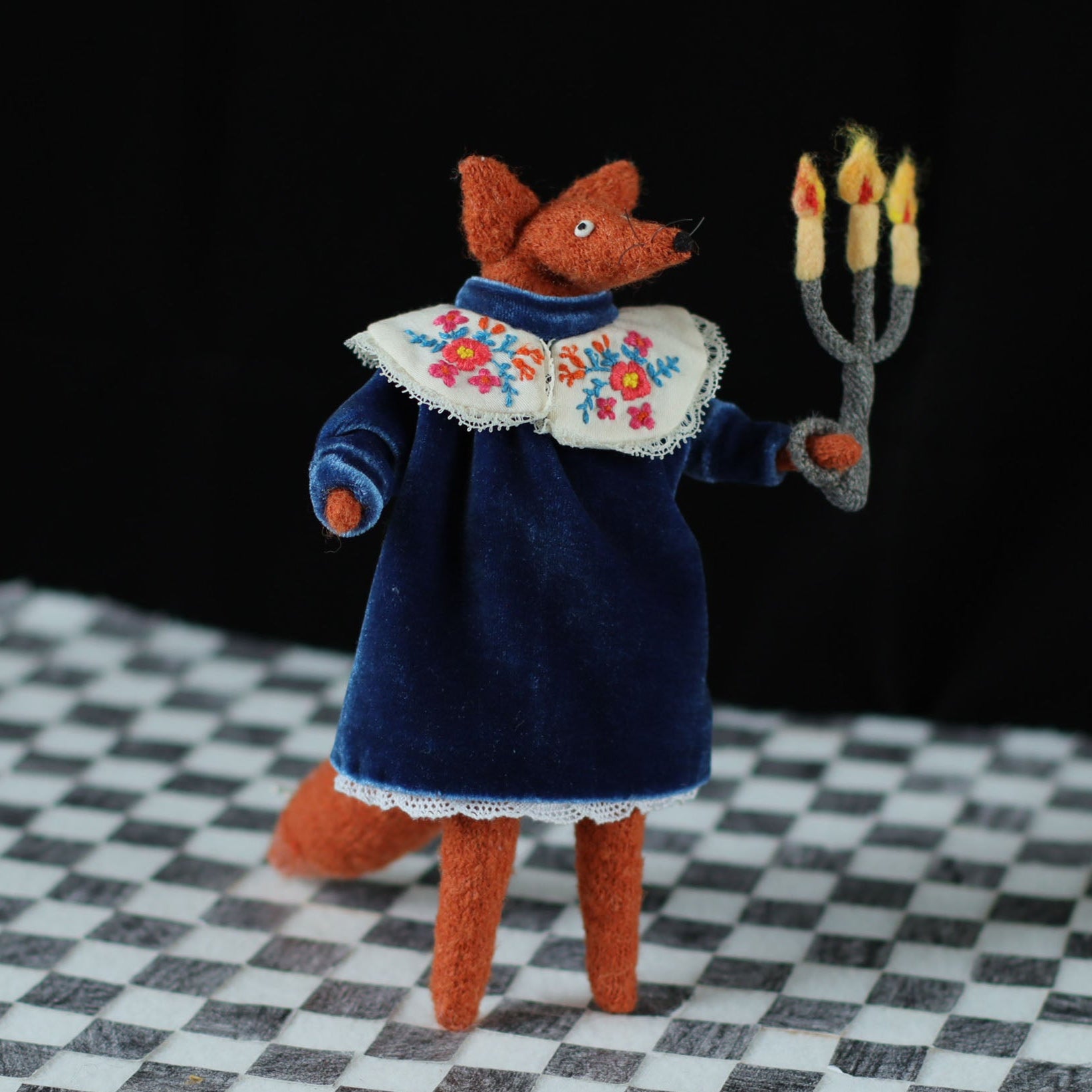 andmade fox doll standing on a checkered floor, dressed in a dark blue velvet dress with an embroidered floral collar, holding a small felt candelabra with glowing yellow-orange flames against a black background