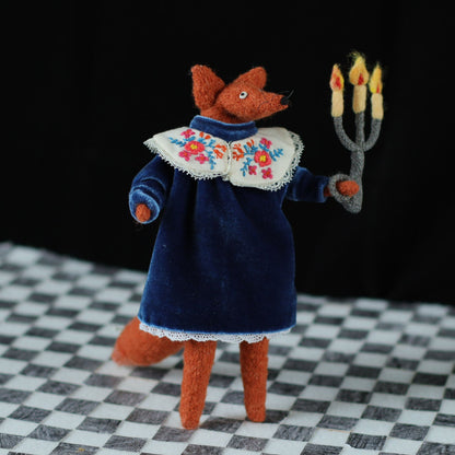 andmade fox doll standing on a checkered floor, dressed in a dark blue velvet dress with an embroidered floral collar, holding a small felt candelabra with glowing yellow-orange flames against a black background