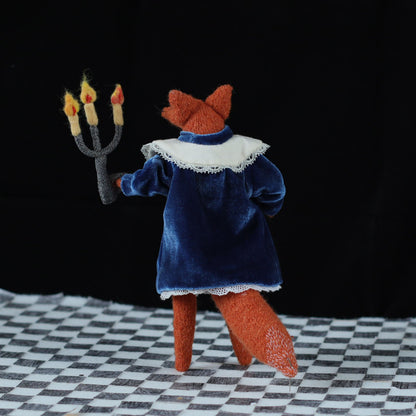 Back view of a handmade fox doll wearing a blue velvet dress with a white lace collar, holding a three-flame felt candelabra while standing on a black-and-white checkered floor against a dark background