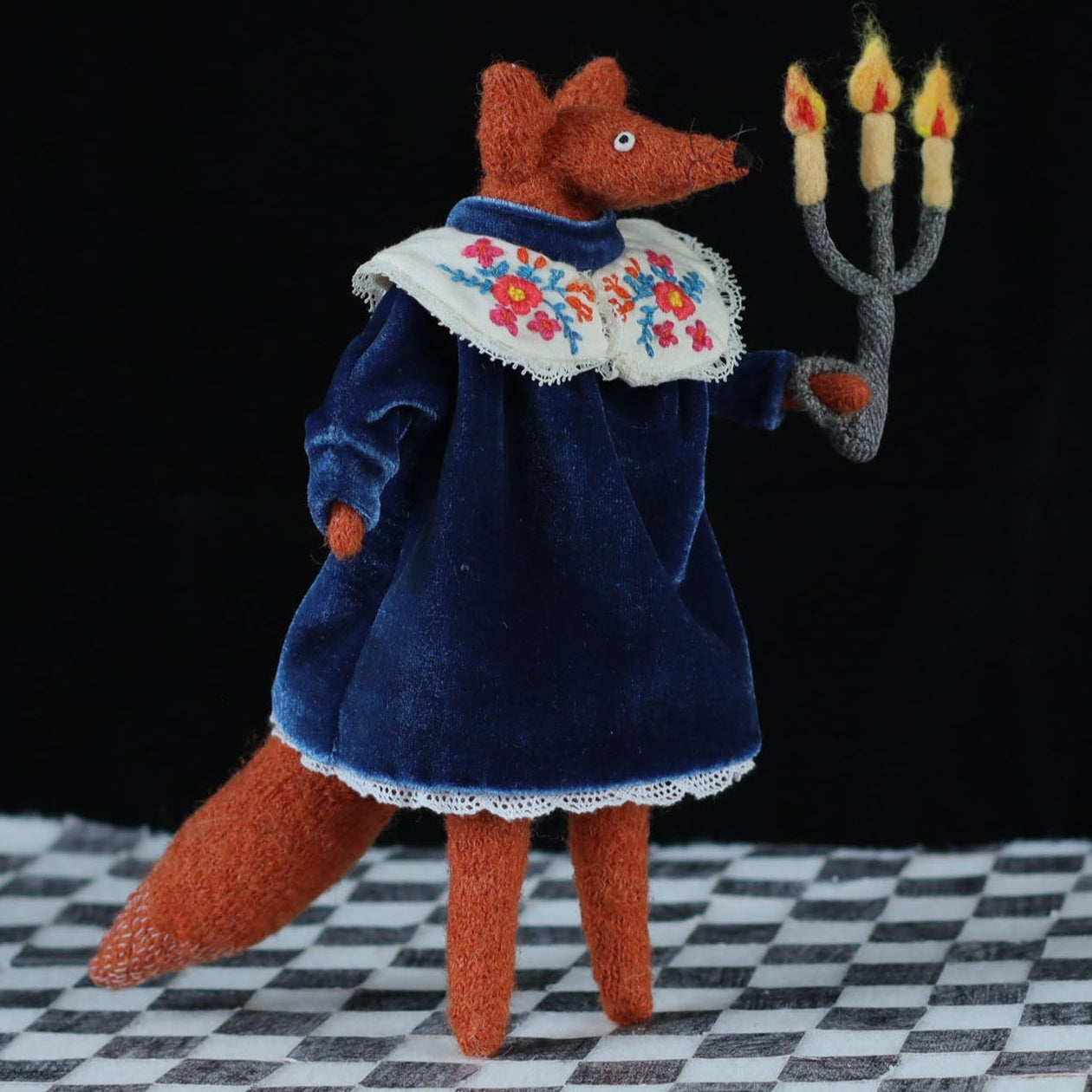 Handmade fox doll standing on a black-and-white checkered floor, dressed in a blue velvet dress with a floral embroidered collar, holding a three-flame felt candelabra against a dark background