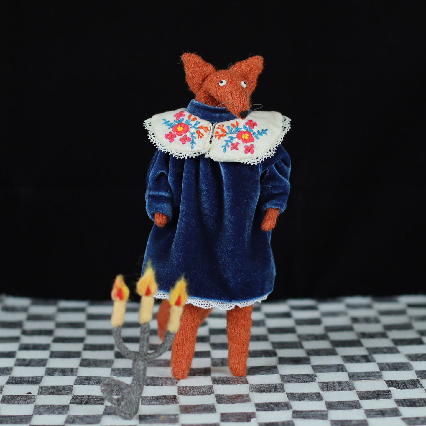 Handmade fox doll in a blue velvet dress with an embroidered floral collar, standing on a checkered floor with a felt candelabra on the floor in front, photographed against a black background