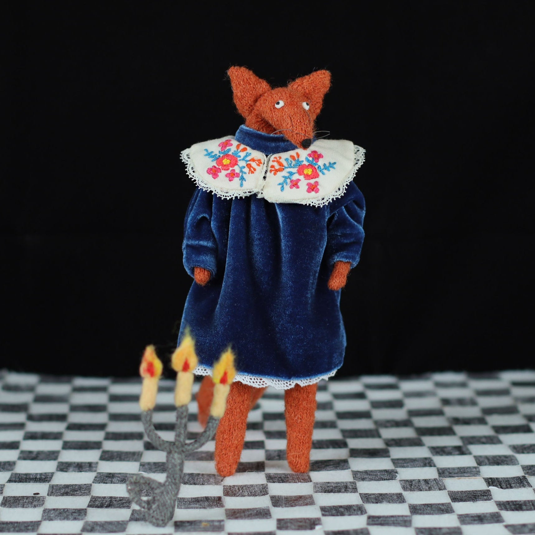 Handmade fox doll in a blue velvet dress with an embroidered floral collar, standing on a checkered floor with a felt candelabra on the floor in front, photographed against a black background
