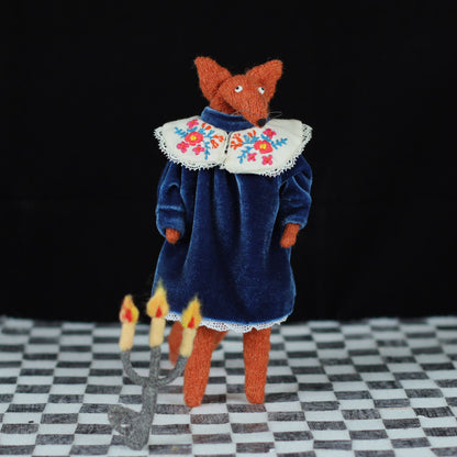 Handmade fox doll in a blue velvet dress with an embroidered floral collar, standing on a checkered floor with a felt candelabra on the floor in front, photographed against a black background