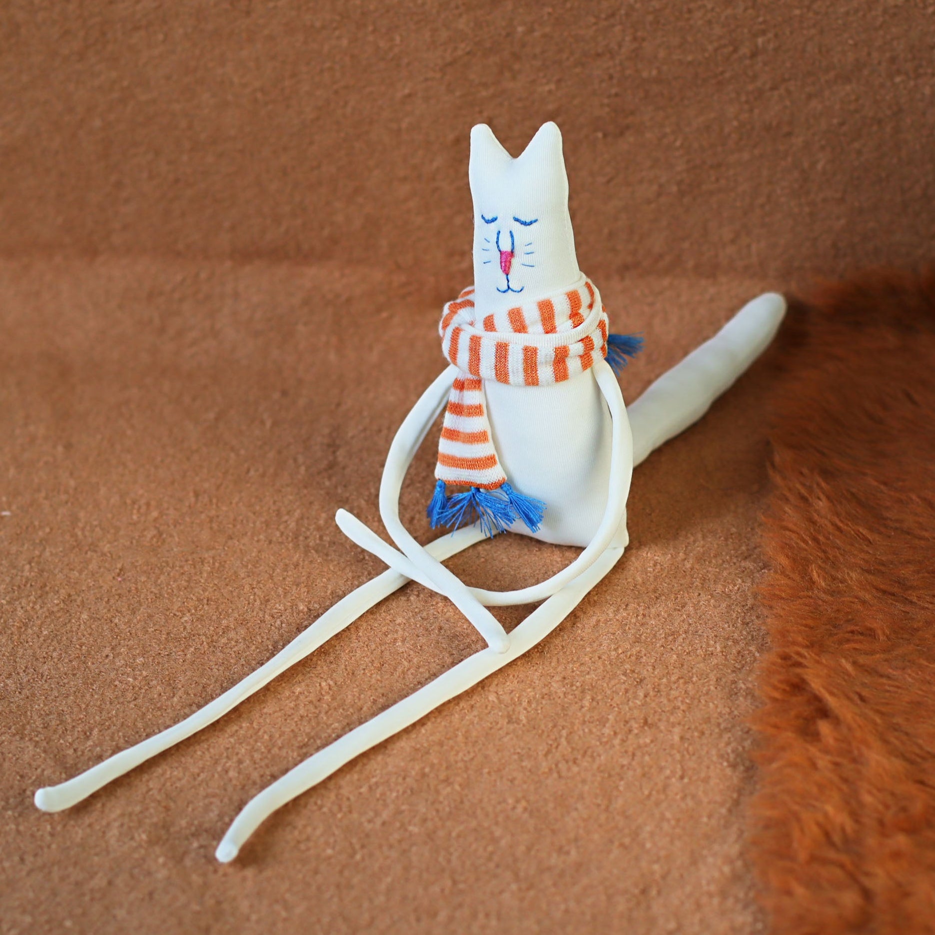 Handmade minimalist cat plush — soft white fabric cat with extra-long limbs and an orange striped scarf, shown seated on a warm brown surface as a collectible art doll