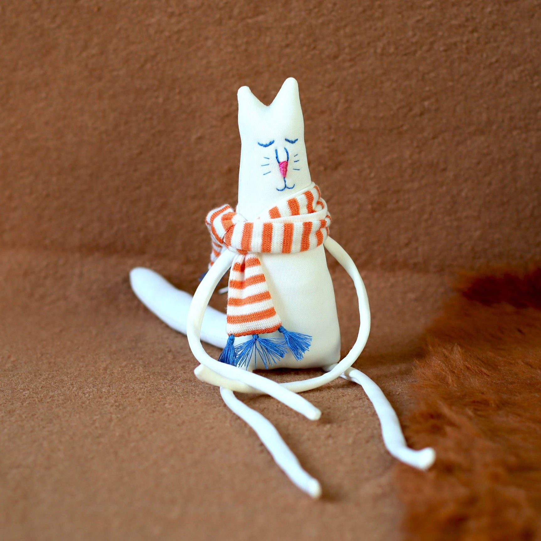 Hand-sewn minimalist white cat plush with extra-long legs, embroidered face, and an orange-and-white striped scarf with blue tassels, seated on a brown background