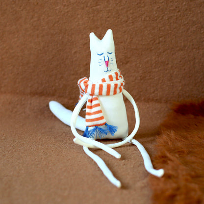 Hand-sewn minimalist white cat plush with extra-long legs, embroidered face, and an orange-and-white striped scarf with blue tassels, seated on a brown background