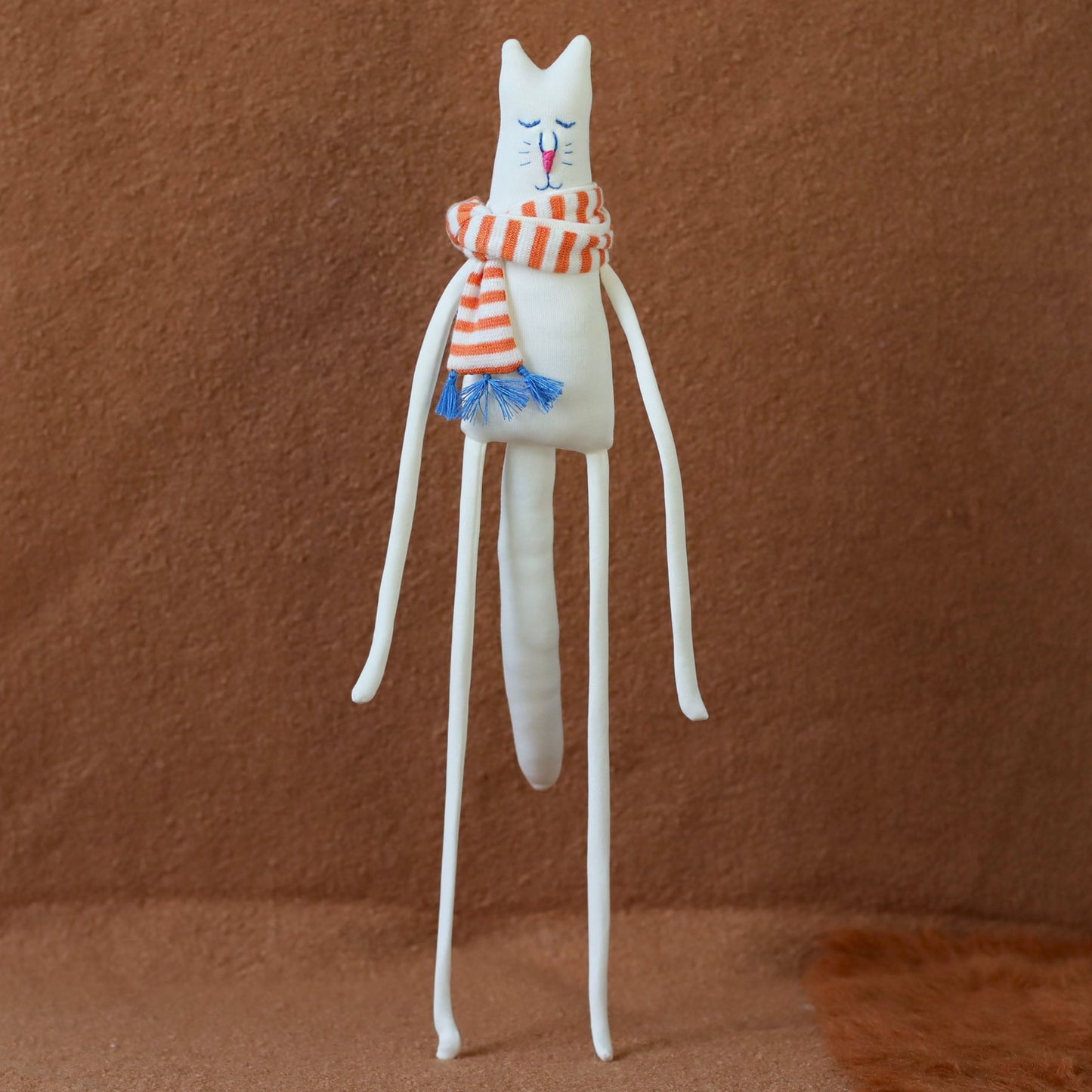 Hand-sewn minimalist white cat plush with long arms and legs, embroidered face, and a red-and-orange striped scarf with blue tassels, standing against a brown background