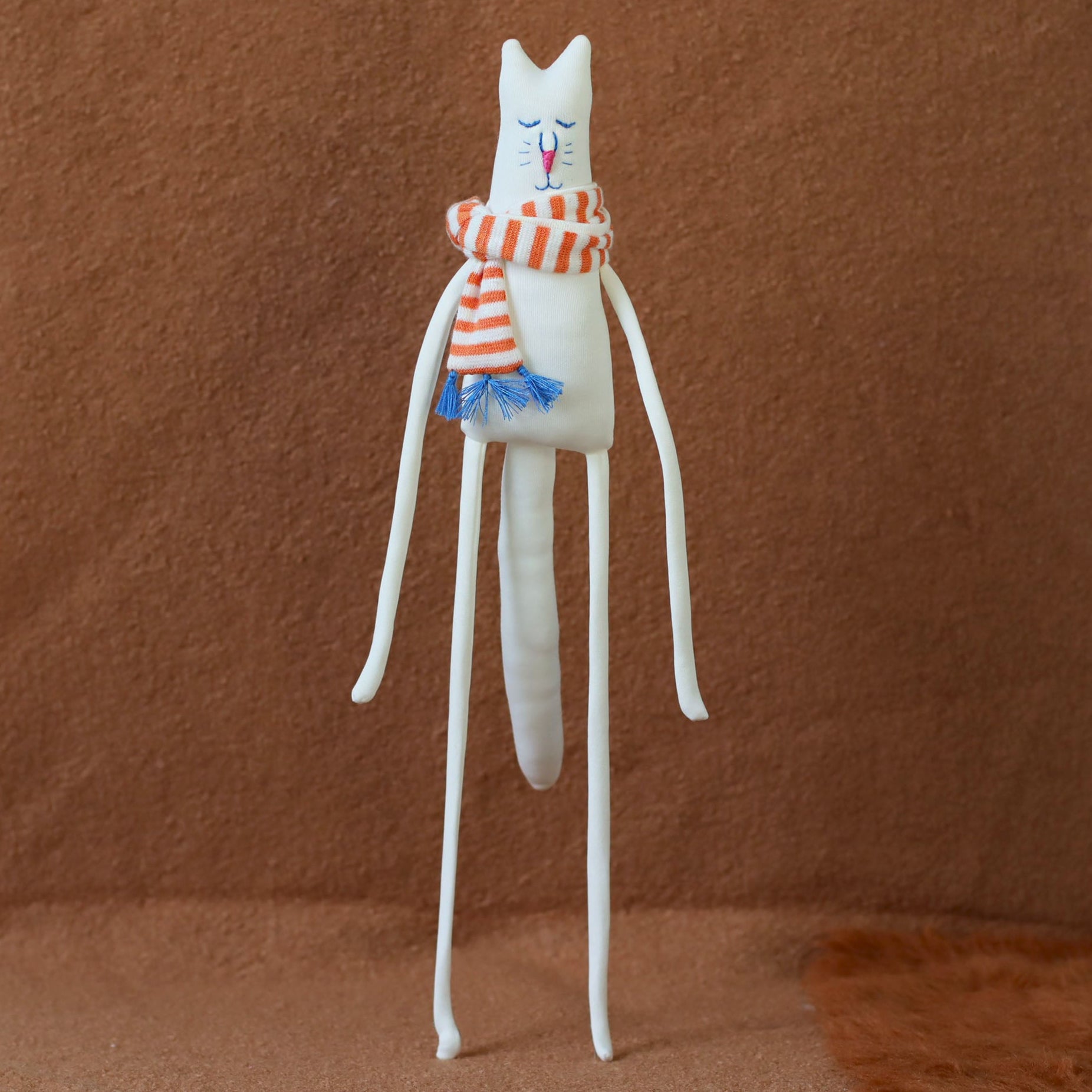 Hand-sewn minimalist white cat plush with long arms and legs, embroidered face, and a red-and-orange striped scarf with blue tassels, standing against a brown background