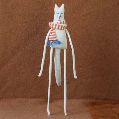 Hand-sewn minimalist white cat plush with long arms and legs, embroidered face, and a red-and-orange striped scarf with blue tassels, standing against a brown background