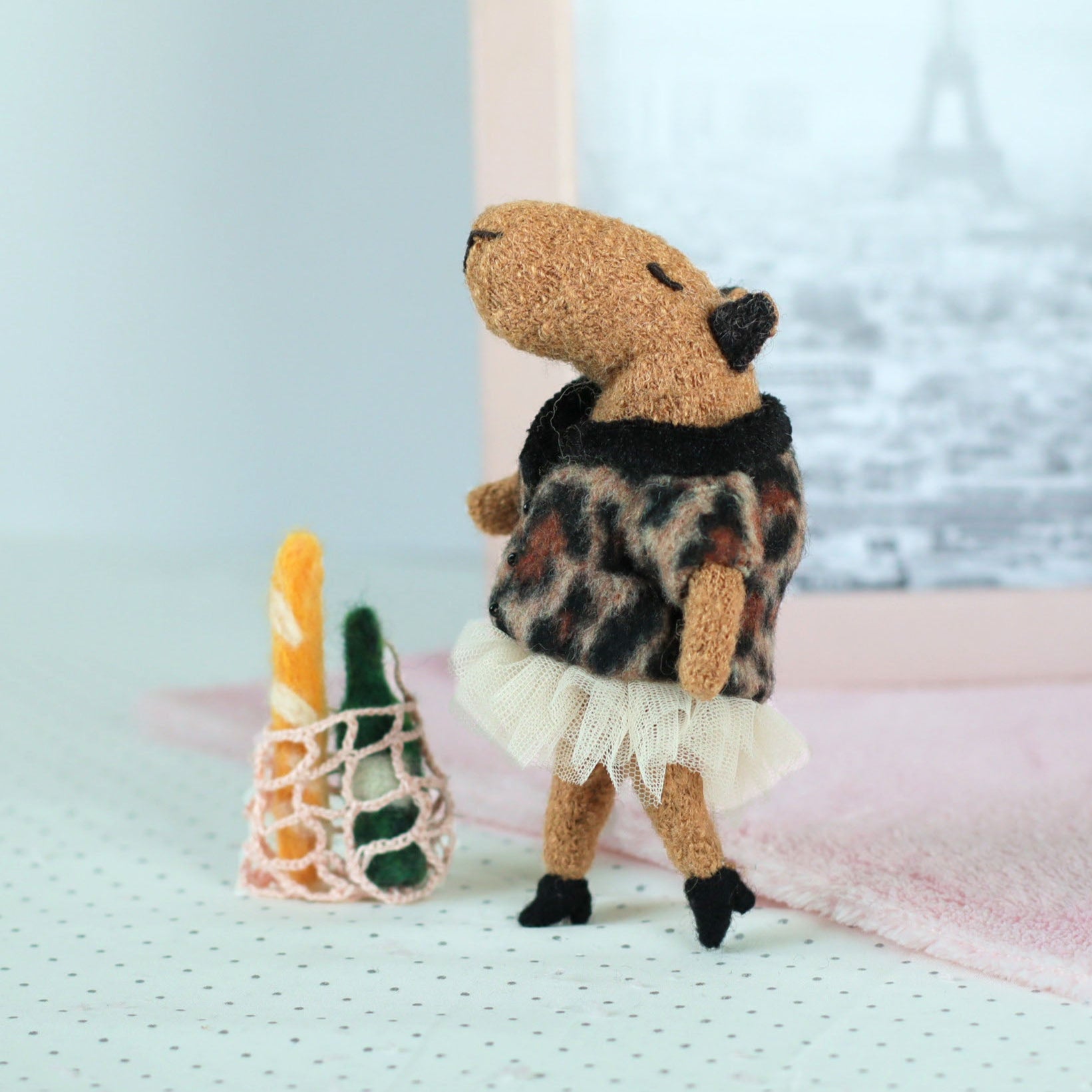 Handmade miniature capybara doll in a leopard-print sweater and tulle skirt standing beside a tiny mesh shopping bag with a felt baguette and bottle, with a soft pink cloth and an Eiffel Tower photo in the background