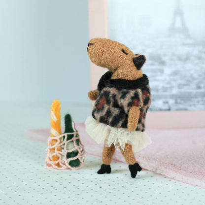Handmade miniature capybara doll in a leopard-print sweater and tulle skirt standing beside a tiny mesh shopping bag with a felt baguette and bottle, with a soft pink cloth and an Eiffel Tower photo in the background