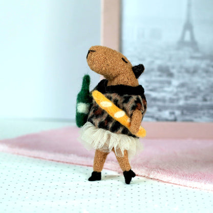 Miniature handmade capybara doll in a leopard-print sweater and tulle skirt, standing and holding a tiny felt baguette and green bottle, with a soft pink cloth and Eiffel Tower photo in the background
