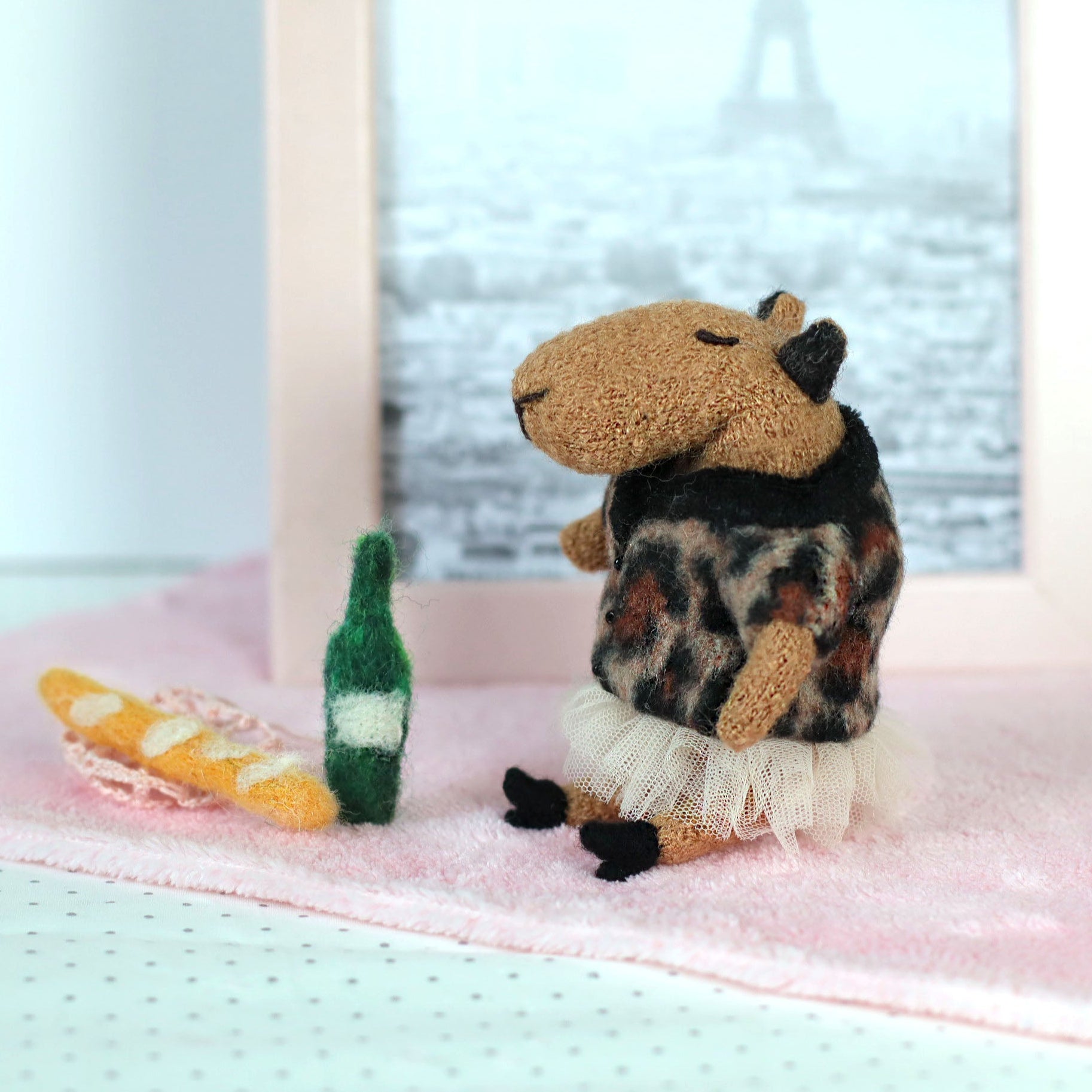 Handmade miniature capybara doll sitting on a pink cloth, wearing a leopard-print sweater and tulle skirt, with tiny felt props including a baguette and green bottle, and a framed Paris photo in the background