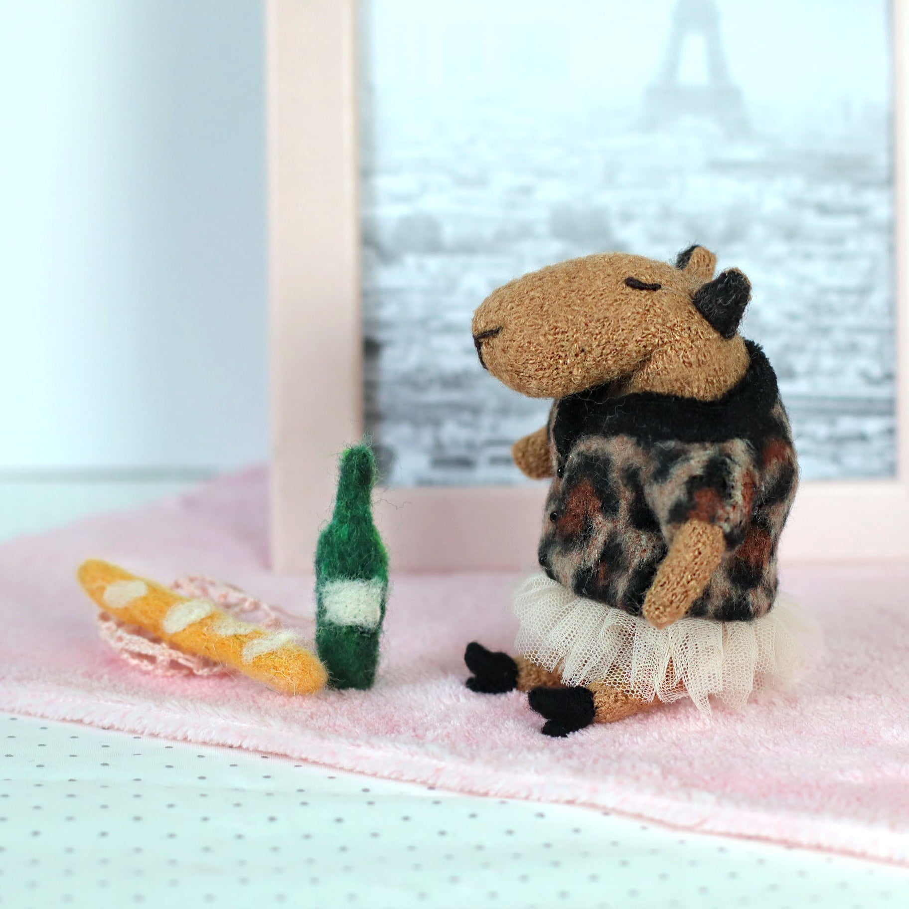 Miniature handmade capybara doll wearing a leopard-print sweater and tulle skirt, sitting on a pink cloth beside a tiny felt baguette and green bottle, with a framed Eiffel Tower photo in the background