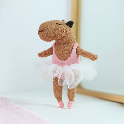 Handmade capybara ballerina doll standing on one leg in front of a mirror, wearing a pink leotard, white tulle tutu, and tiny pink ballet shoes